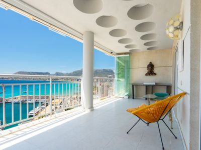 Vaste terrasse couverte avec carrelage blanc et plafond à motifs circulaires. Offre des vues panoramiques sur le port de plaisance, la mer turquoise et les montagnes lointaines, avec parois vitrées.
