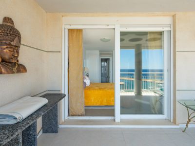 Vue de la terrasse vers une chambre avec un couvre-lit jaune vif, agrémentée d'un buste en bois sculpté et d'une console en granit. Les portes coulissantes en verre offrent un aperçu de la mer.