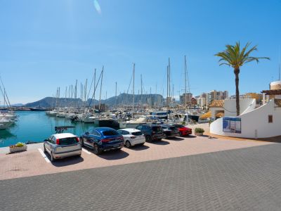 Vue du port de plaisance de Calpe avec de nombreux voiliers, voitures garées et bâtiments côtiers sous un ciel bleu clair, encadrée par un palmier.