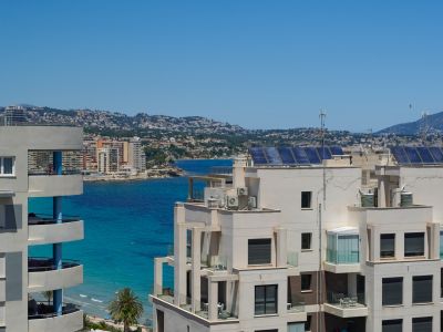 Vue élevée sur des bâtiments côtiers modernes avec balcons, surplombant la mer turquoise, la côte lointaine et les montagnes sous un ciel clair. Panneaux solaires visibles sur les toits.