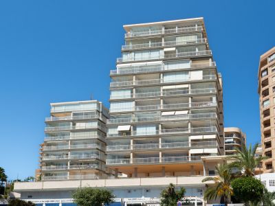 Vue extérieure d'un immeuble résidentiel moderne à plusieurs étages avec de nombreux balcons, dont plusieurs sont fermés par des rideaux de verre et des balustrades blanches, sous un ciel bleu. Des palmiers sont visibles.