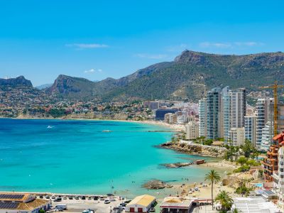 Vue panoramique de la baie de Calpe, avec ses eaux turquoise, plages de sable, immeubles côtiers modernes et montagnes verdoyantes sous ciel bleu.