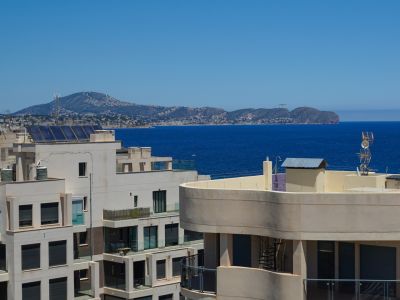 Vue panoramique sur la mer depuis un étage élevé, avec architecture moderne adjacente, panneaux solaires et une côte montagneuse urbanisée lointaine.