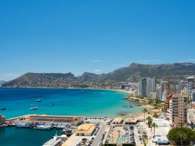 Vue panoramique sur le port de plaisance de Calpe, la baie turquoise et la Playa de La Fossa, encadrée par des montagnes lointaines sous un ciel clair.