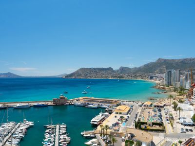 Vue panoramique sur le port de plaisance de Calpe, sa baie aux eaux turquoise, une plage de sable bordée de bâtiments côtiers et des montagnes lointaines.