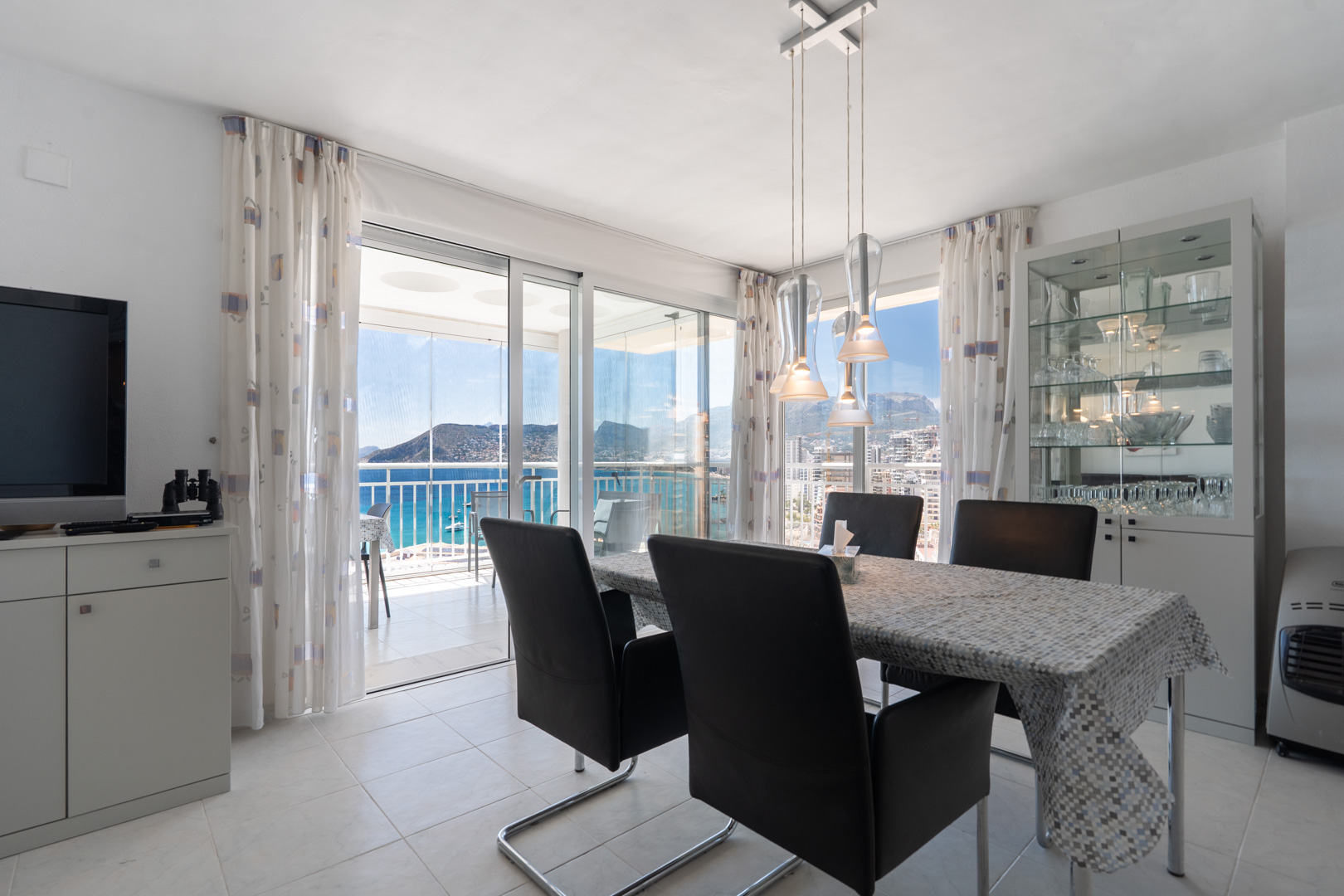 Salle à manger lumineuse avec mobilier moderne et luminaire suspendu. Grandes portes vitrées s'ouvrent sur une terrasse couverte, offrant des vues panoramiques sur la mer et la montagne.