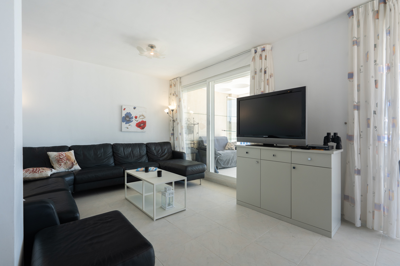 Salon lumineux avec canapé d'angle en cuir noir, table basse blanche et TV sur meuble gris. Des portes coulissantes mènent à une terrasse couverte.