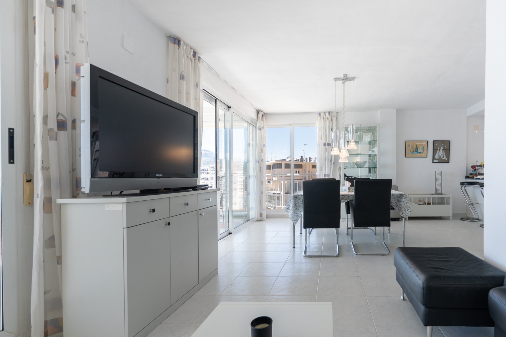 Séjour-salle à manger lumineux avec carrelage blanc, télévision sur meuble gris et table à manger avec suspensions modernes. Les baies vitrées s'ouvrent sur un balcon offrant des vues urbaines.