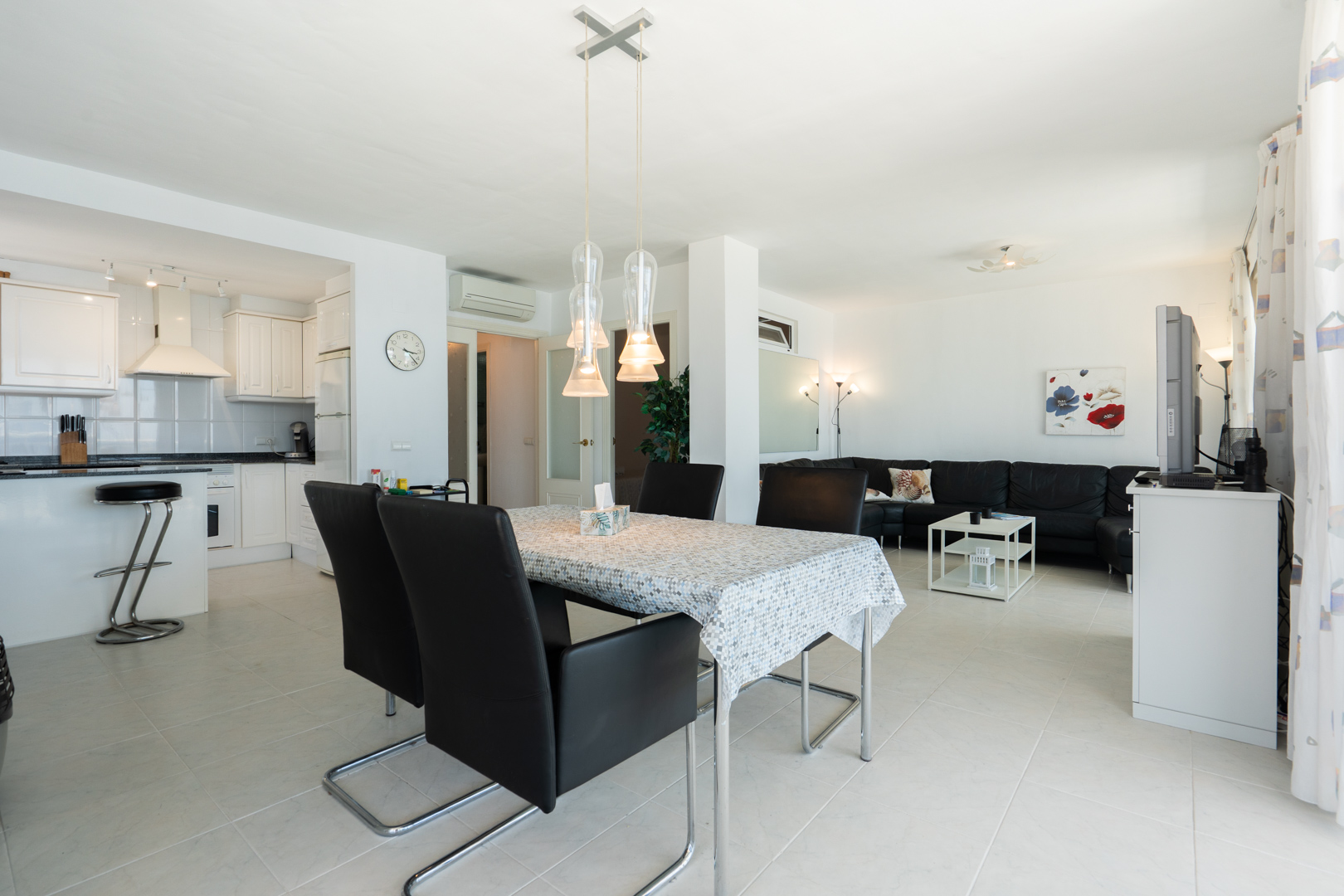 Vaste espace de vie ouvert comprenant une table à manger avec des suspensions modernes, une cuisine blanche intégrée avec bar et un salon confortable avec canapé sectionnel noir.