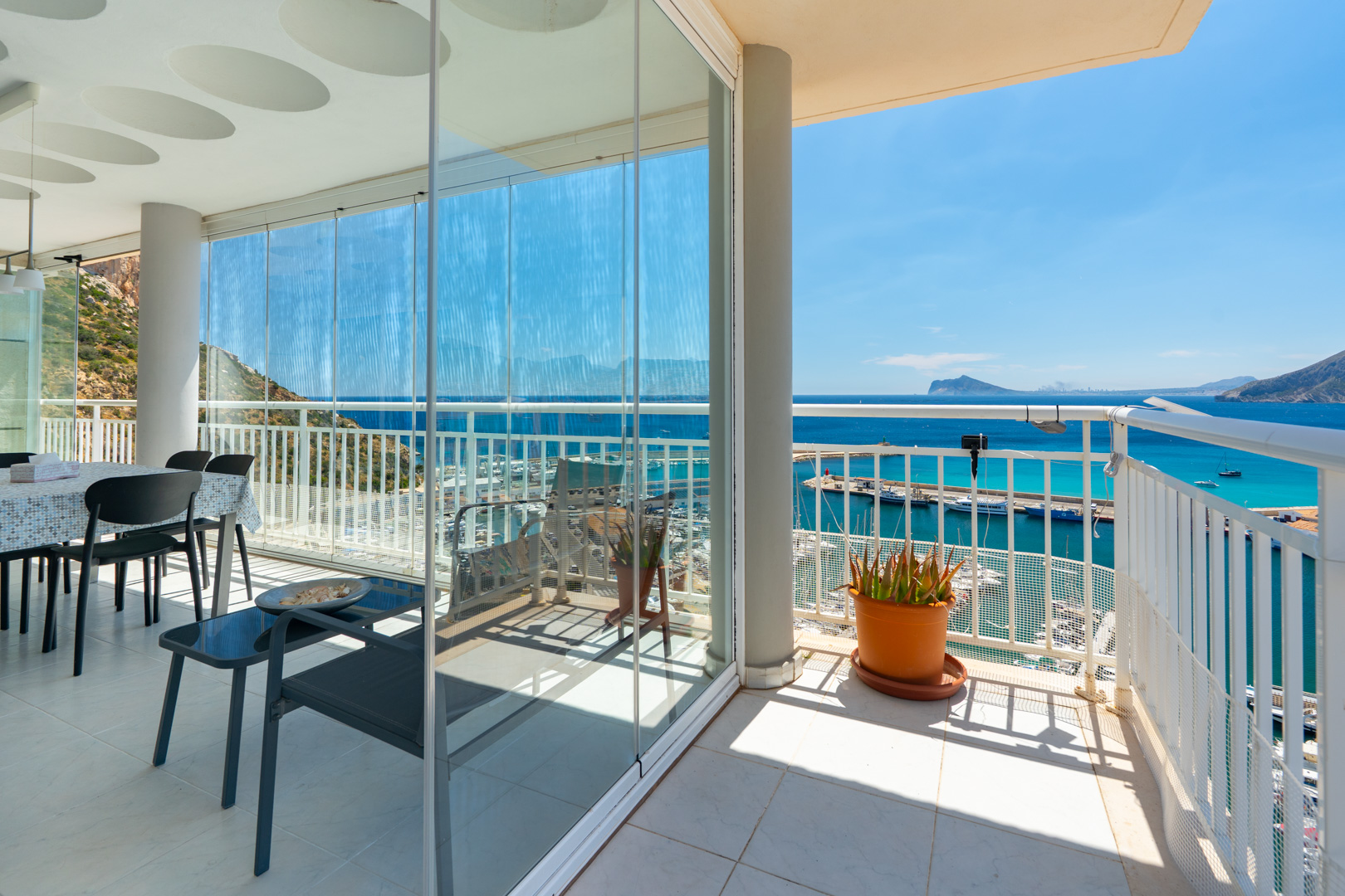 Vaste terrasse avec rideaux de verre délimitant un espace repas, s'ouvrant sur un balcon. Offre des vues panoramiques sur le port de Calpe, la mer azur et le littoral lointain sous un ciel clair.