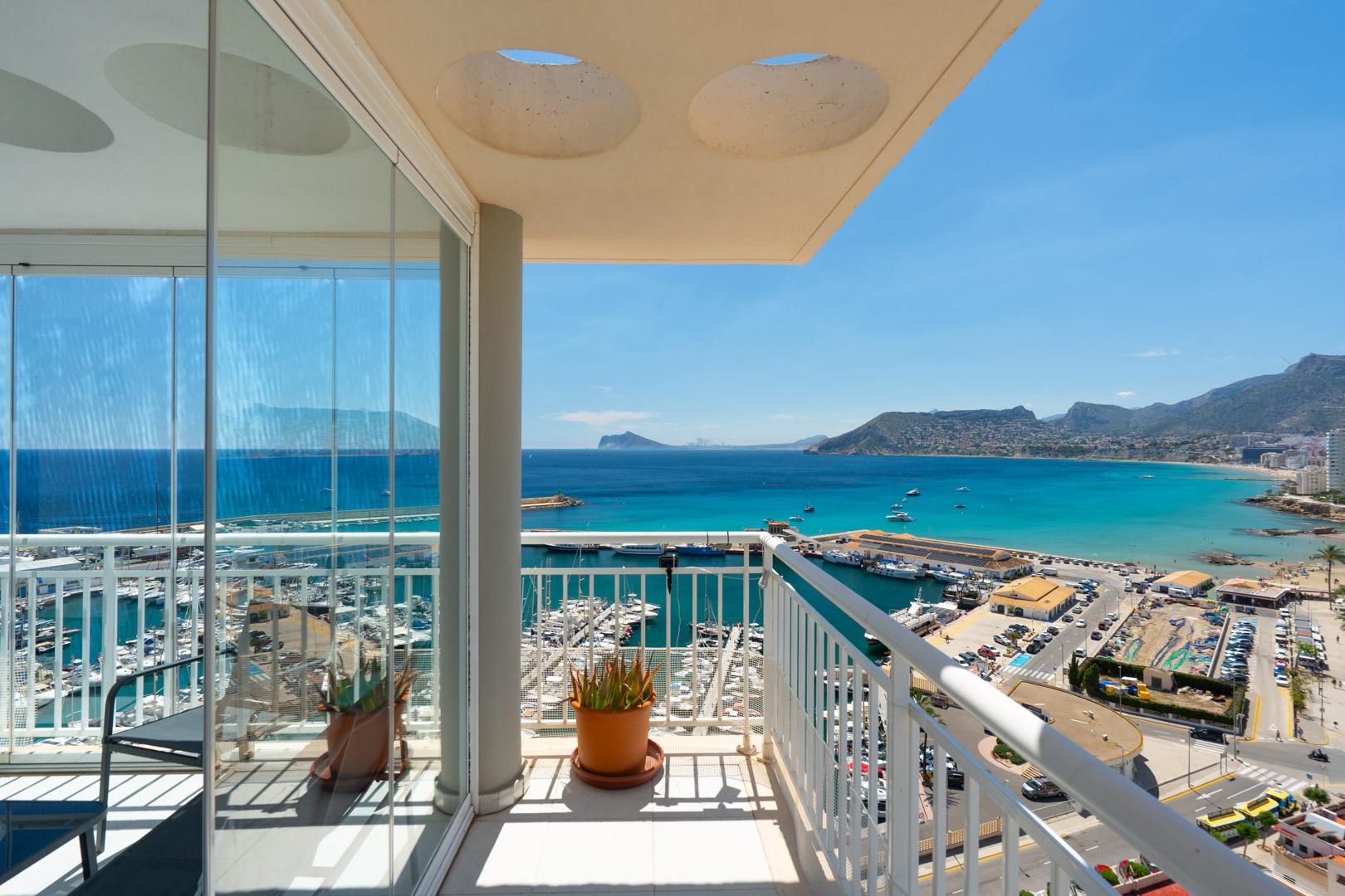Vaste terrasse offrant une vue panoramique sur le port de plaisance de Calpe, la baie turquoise et le littoral lointain, encadrée de rideaux de verre et d'un garde-corps blanc.