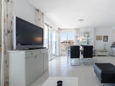 Séjour-salle à manger lumineux avec carrelage blanc, télévision sur meuble gris et table à manger avec suspensions modernes. Les baies vitrées s'ouvrent sur un balcon offrant des vues urbaines.