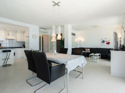Vaste espace de vie ouvert comprenant une table à manger avec des suspensions modernes, une cuisine blanche intégrée avec bar et un salon confortable avec canapé sectionnel noir.