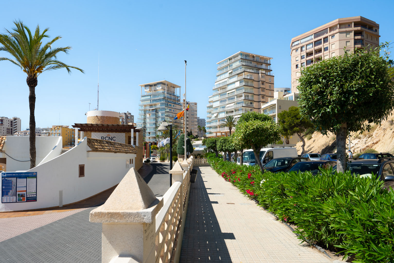 Allée piétonne pavée avec balustrade en pierre et lauriers-roses luxuriants, menant vers des immeubles modernes et l'entrée du Real Club Náutico Calpe sous un ciel bleu clair.