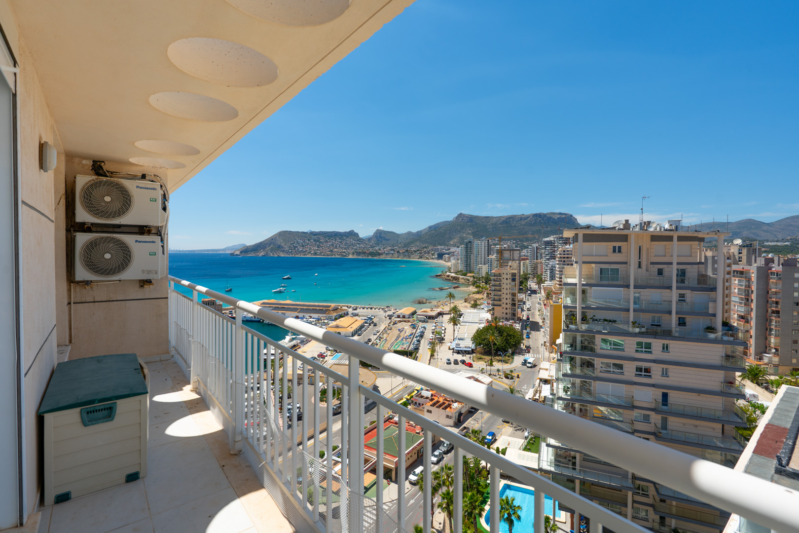 Balcon offrant une vue panoramique sur le port de Calpe, la baie turquoise et le littoral urbain, encadré par une balustrade blanche et des unités de climatisation extérieures.