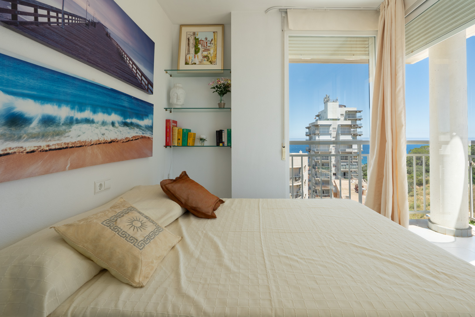 Chambre à coucher avec lit confortable, décorée de tableaux marins et étagères en verre. Accès à un balcon offrant des vues partielles sur la mer et la ville.