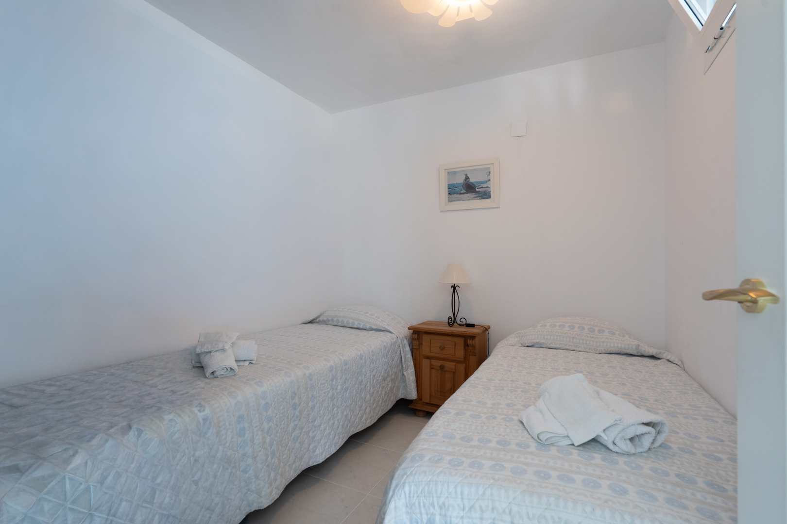 Chambre lumineuse avec deux lits simples aux parures claires, une table de chevet en bois avec lampe, un tableau décoratif et une fenêtre en hauteur.