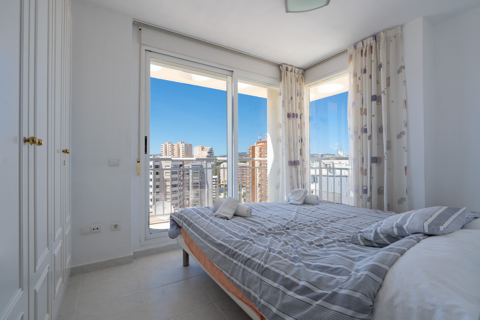 Chambre lumineuse avec lit double, armoire intégrée et portes coulissantes vers une terrasse. Vues urbaines dégagées sous un ciel bleu.