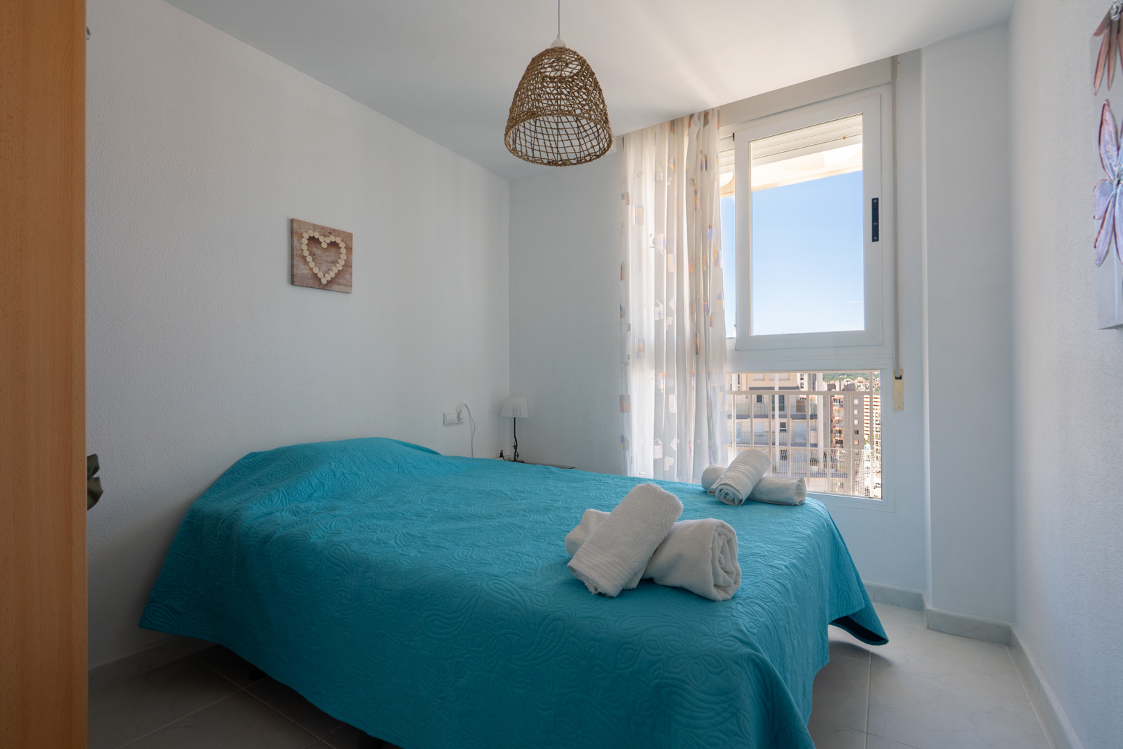 Chambre lumineuse avec lit double en parure turquoise, suspension en fibres naturelles et fenêtre offrant une vue urbaine sur les bâtiments environnants.