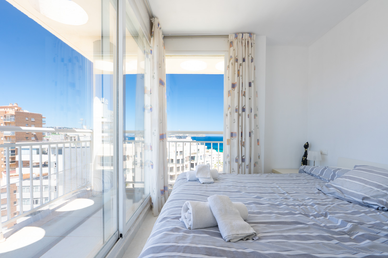 Chambre lumineuse avec lit double et linge de lit rayé, offrant un accès direct à une terrasse avec vue panoramique sur la mer et le ciel.