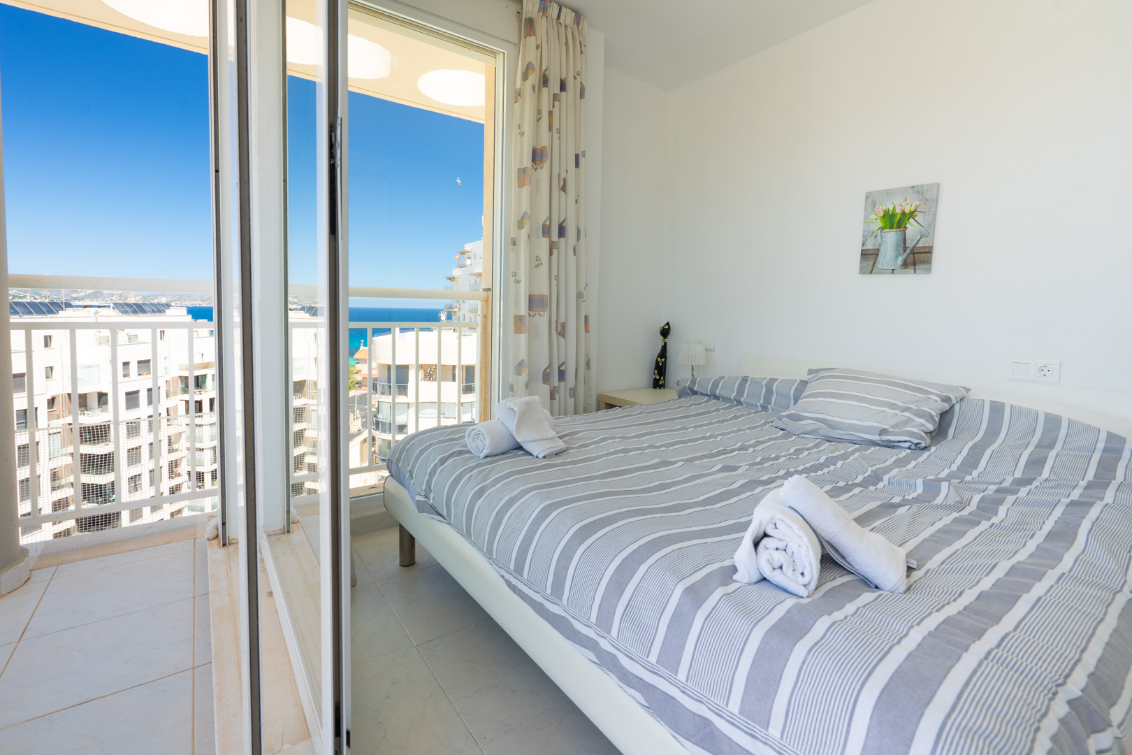 Chambre lumineuse avec lit double, linge de lit rayé, lampe de chevet et accès direct à un balcon privé offrant vue sur mer et ville.