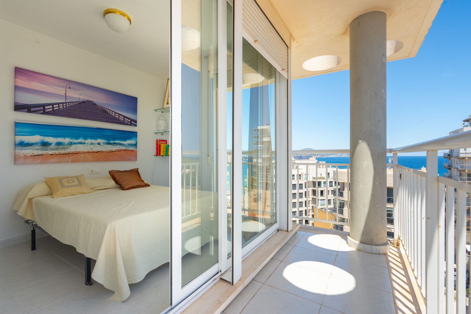 Chambre lumineuse avec lit simple et art mural marin. Accès direct à un balcon privé avec balustrade blanche, pilier en béton et vues sur la mer et la côte.