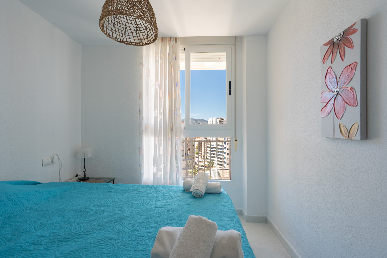 Chambre lumineuse avec un lit au couvre-lit bleu matelassé et serviettes blanches. Fenêtre offrant des vues urbaines et montagneuses, suspension en osier et tableau floral.