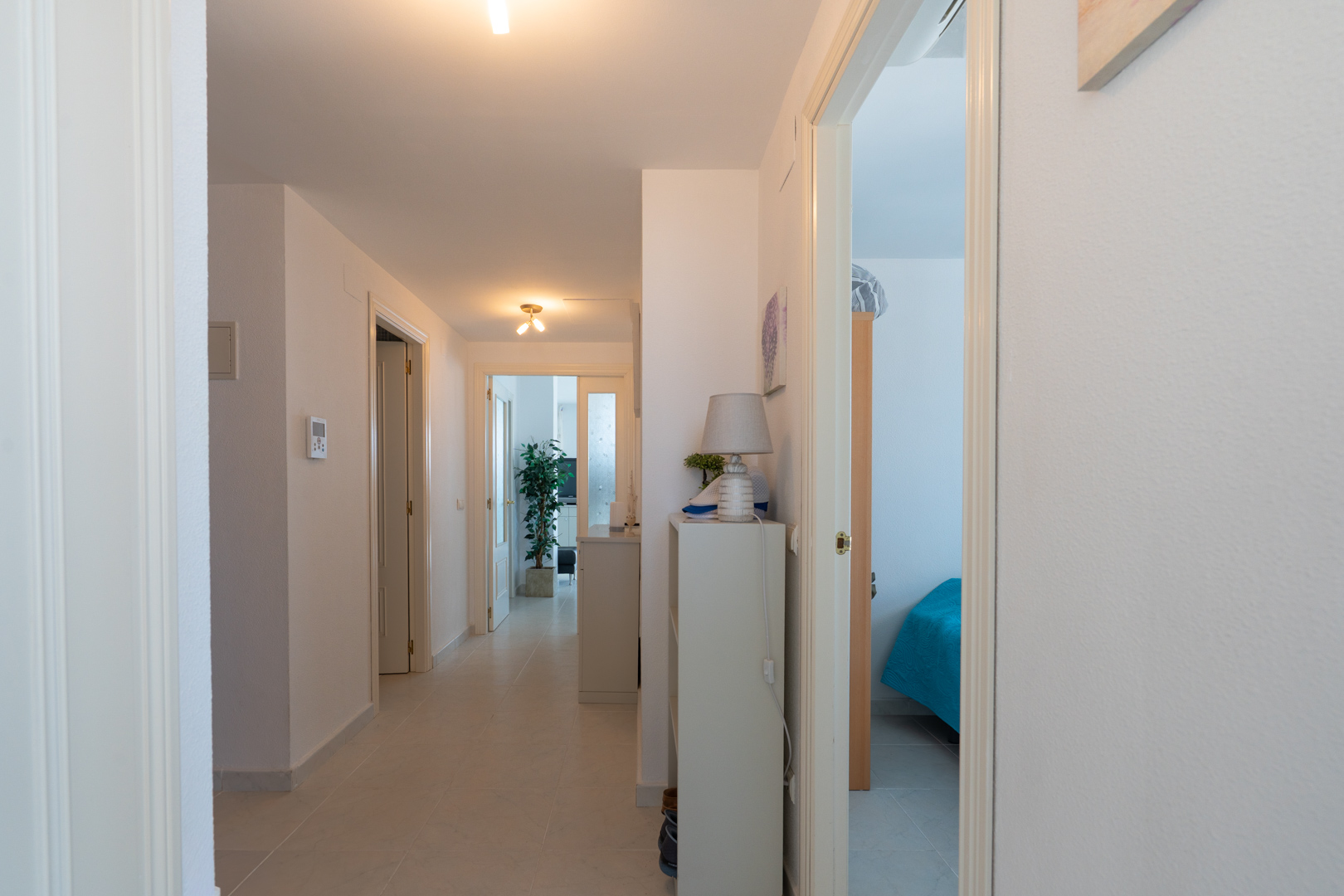 Couloir lumineux avec carrelage clair, portes blanches et une console avec lampe. Aperçu d'une chambre avec lit bleu et d'une autre pièce avec plante.