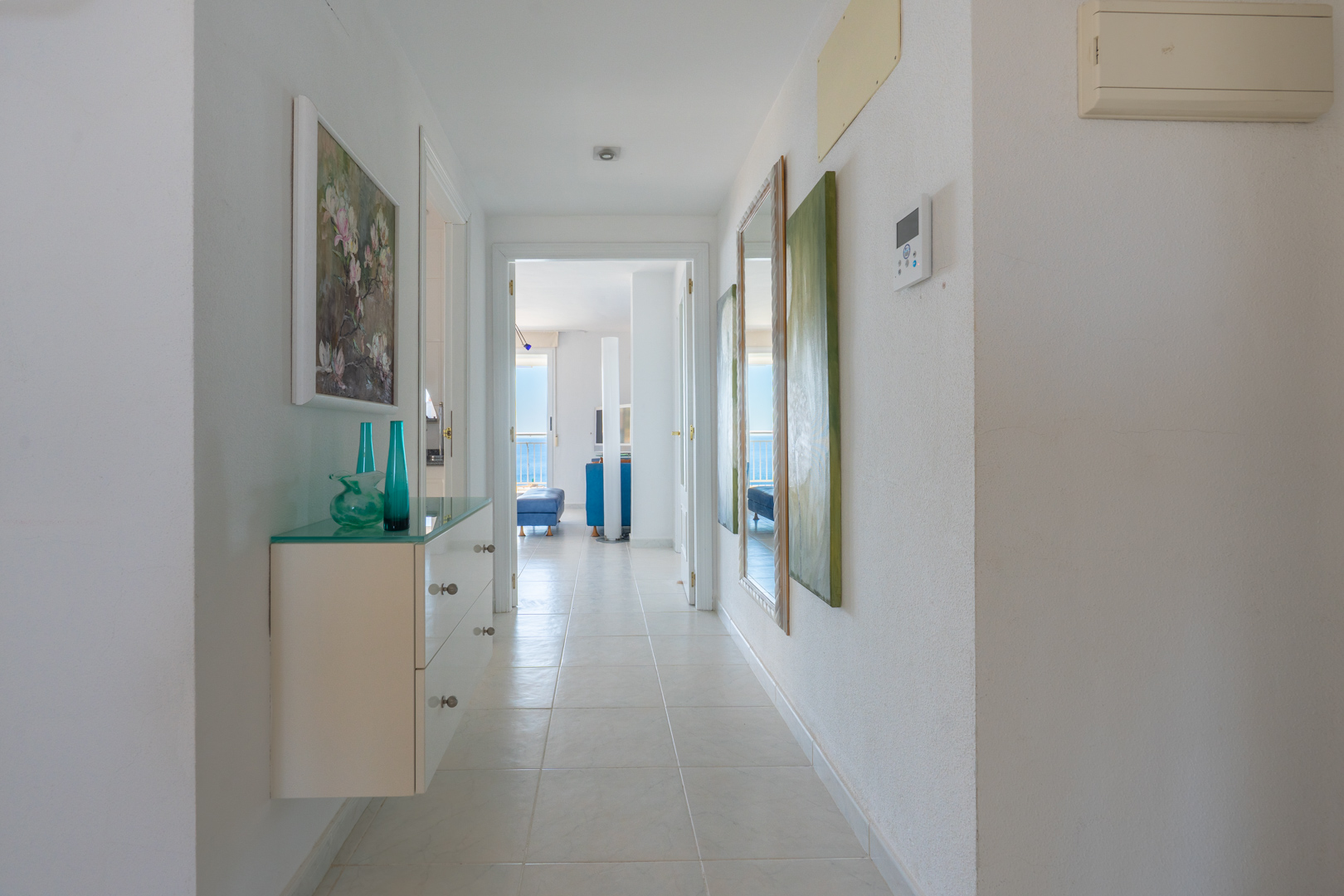 Couloir lumineux avec console flottante en verre, vases décoratifs et tableau floral. Il mène à une pièce principale offrant une vue sur la mer.
