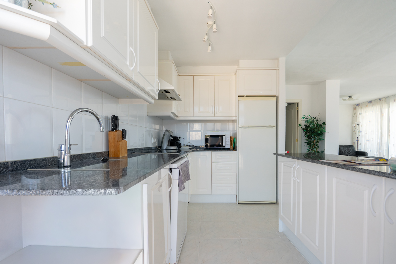 Cuisine fonctionnelle avec des armoires blanches, des plans de travail en granit foncé et un carrelage mural blanc. Équipée d'appareils essentiels et d'un bar ouvrant sur le salon.