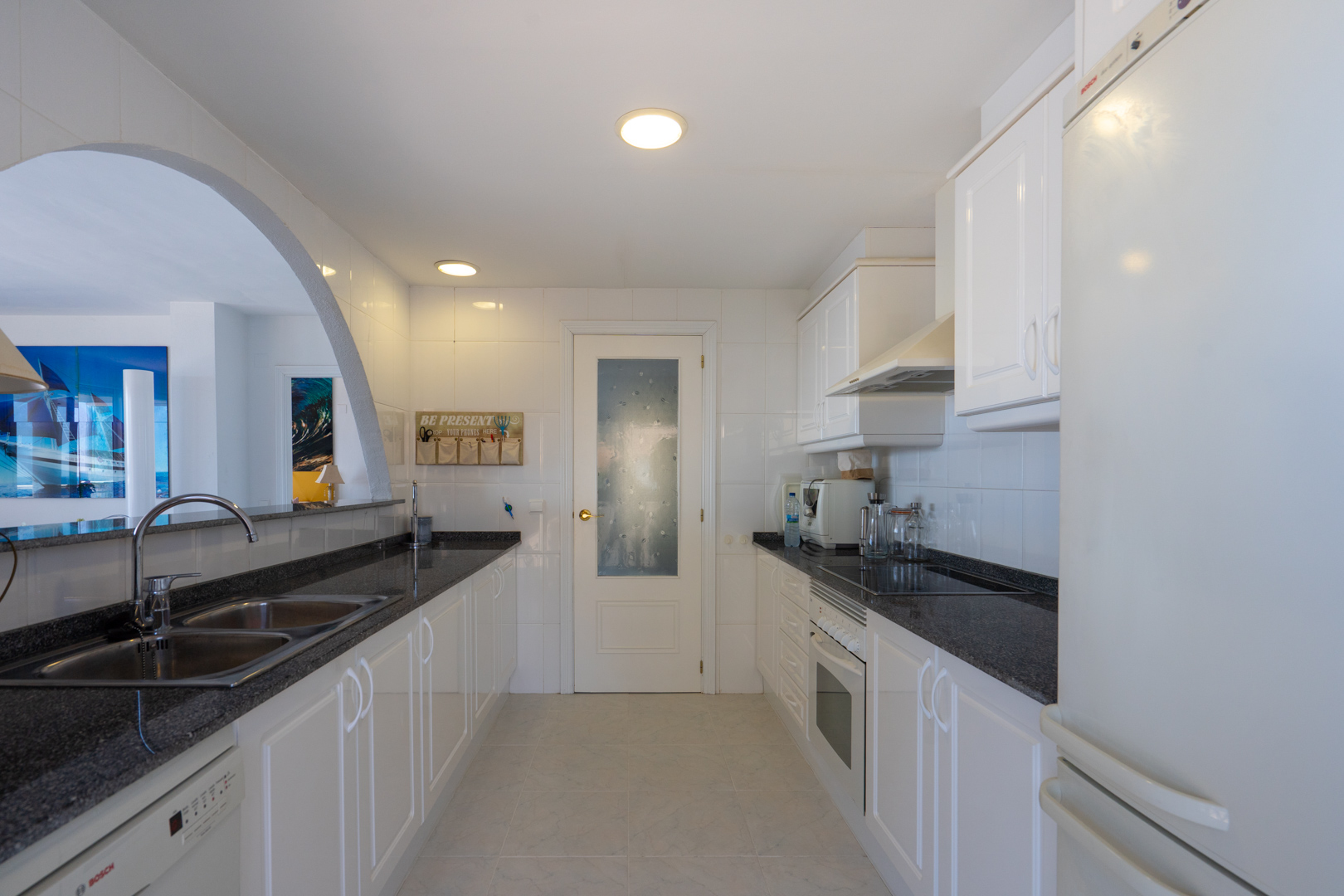 Cuisine fonctionnelle avec meubles blancs, plans de travail en granit foncé et appareils intégrés, reliée au salon par une arche.