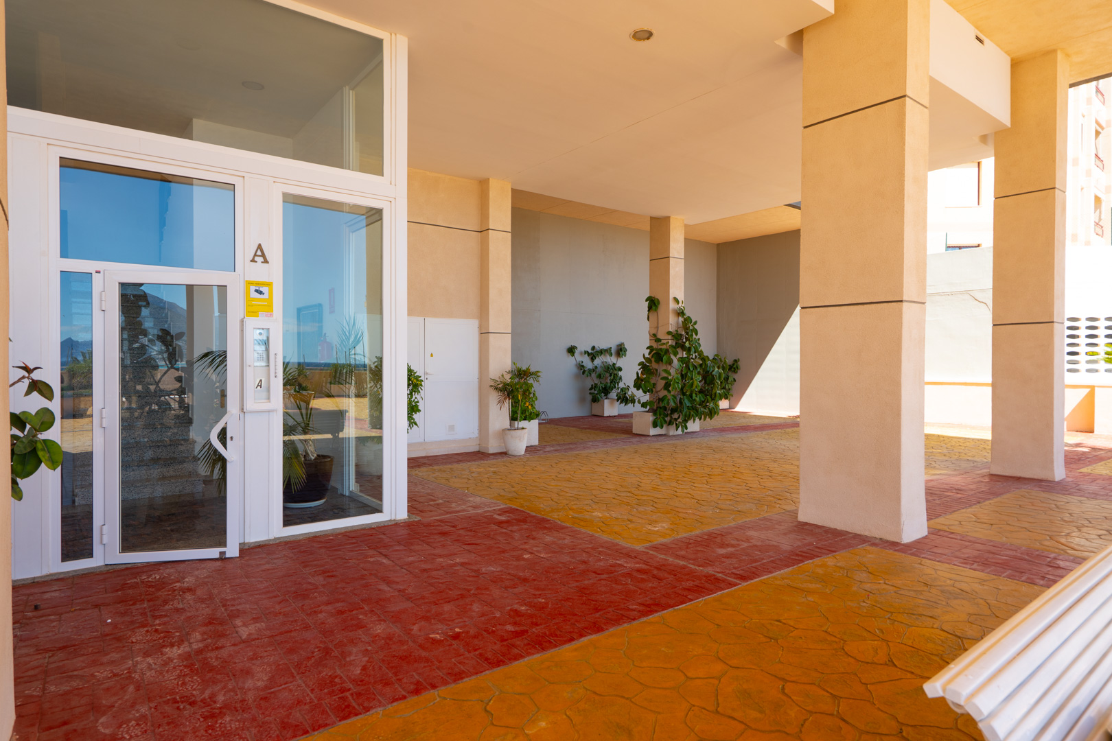 Entrée principale avec portes vitrées à cadre blanc, sol pavé bicolore rouge et orange, et piliers de soutien beiges. Des plantes en pot agrémentent cet espace extérieur couvert.
