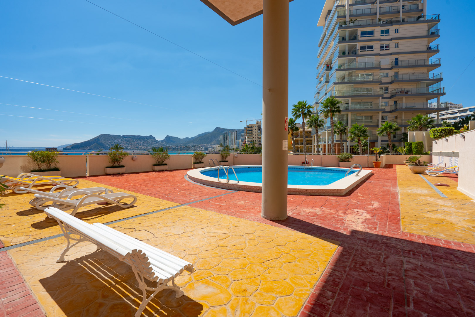 Espace piscine commun avec dallage bicolore rouge et jaune, équipé de transats. Vue sur la mer, les montagnes et un immeuble résidentiel moderne.