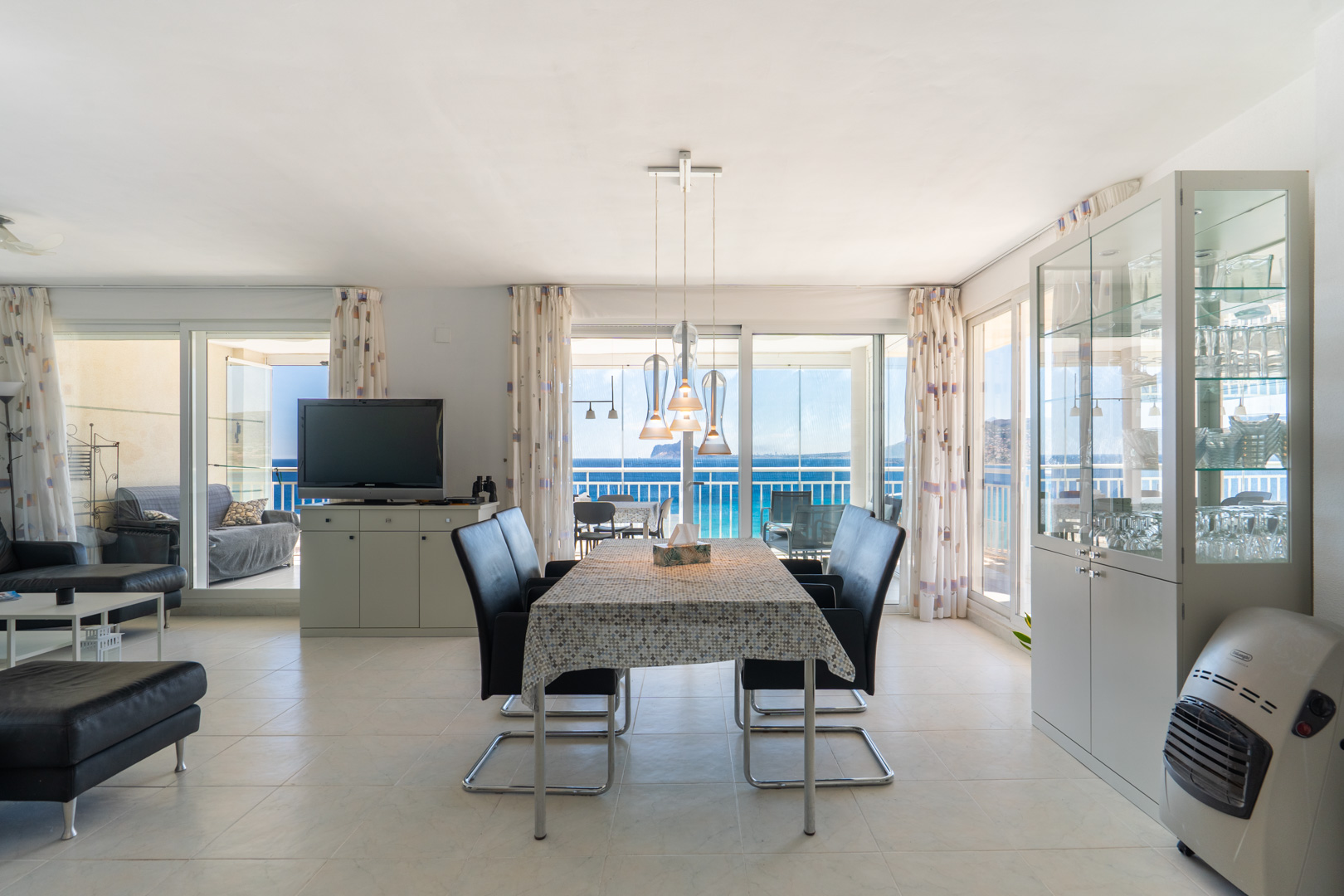 Grand espace de vie et salle à manger ouvert avec carrelage clair, comprenant une table moderne et des vues directes sur la mer par de larges baies vitrées menant à une terrasse vitrée.