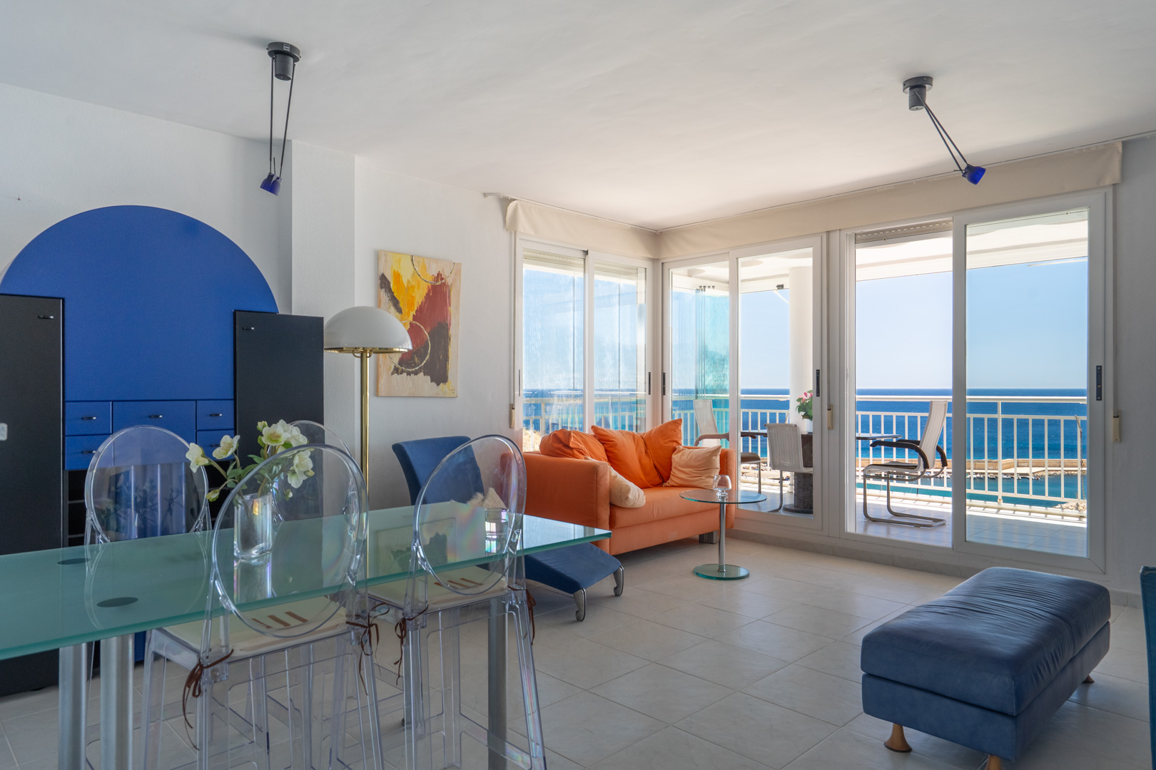 Grand salon-salle à manger avec canapé orange, table en verre et chaises transparentes. De larges baies vitrées s'ouvrent sur une terrasse offrant une vue directe sur la mer.