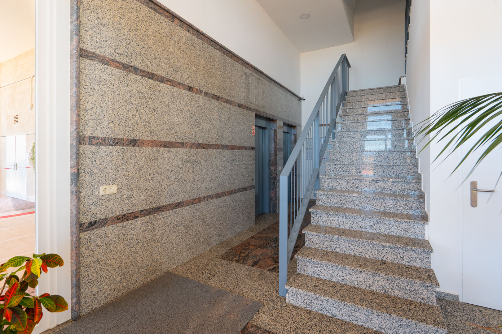 Hall d'entrée avec sol et escalier en granit poli et rampe métallique grise. Murs revêtus de pierre mouchetée avec bandes décoratives, à côté de deux ascenseurs, éclairé par la lumière naturelle.