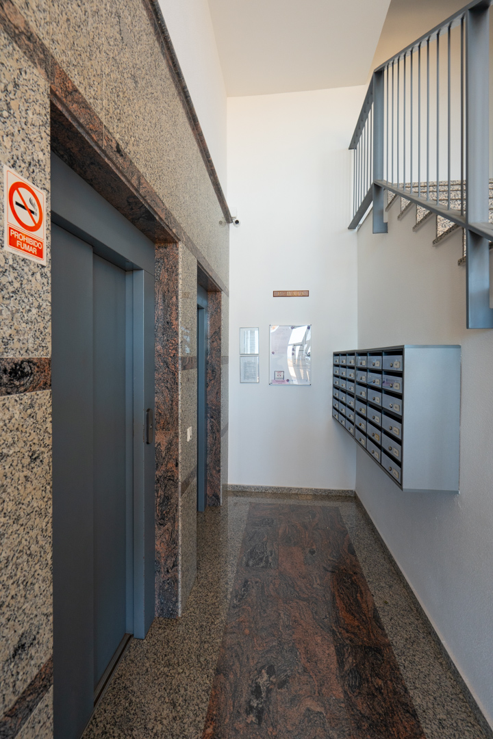 Hall d'entrée de l'immeuble avec deux ascenseurs encadrés de granit moucheté, bande de sol en granit foncé contrastant, boîtes aux lettres communes et escalier métallique.