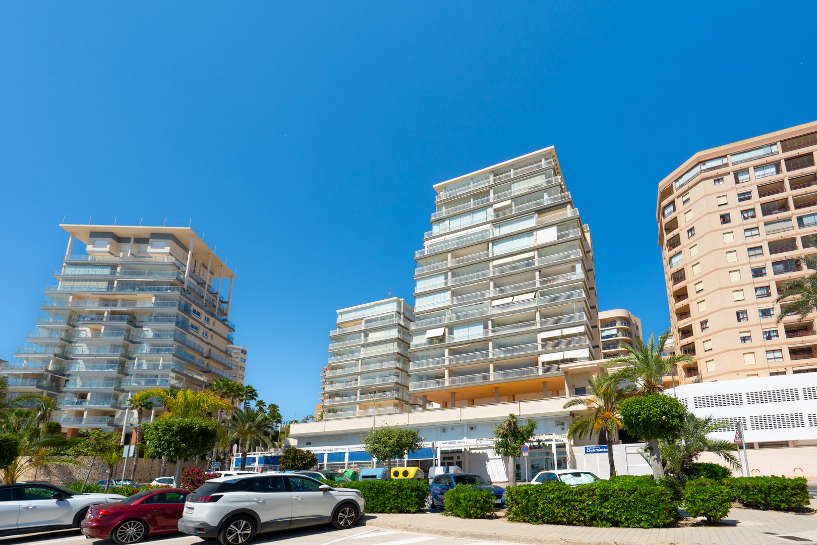 Immeubles résidentiels modernes avec balcons vitrés et un bâtiment à façade traditionnelle, bordant une rue avec palmiers et véhicules stationnés sous un ciel bleu.