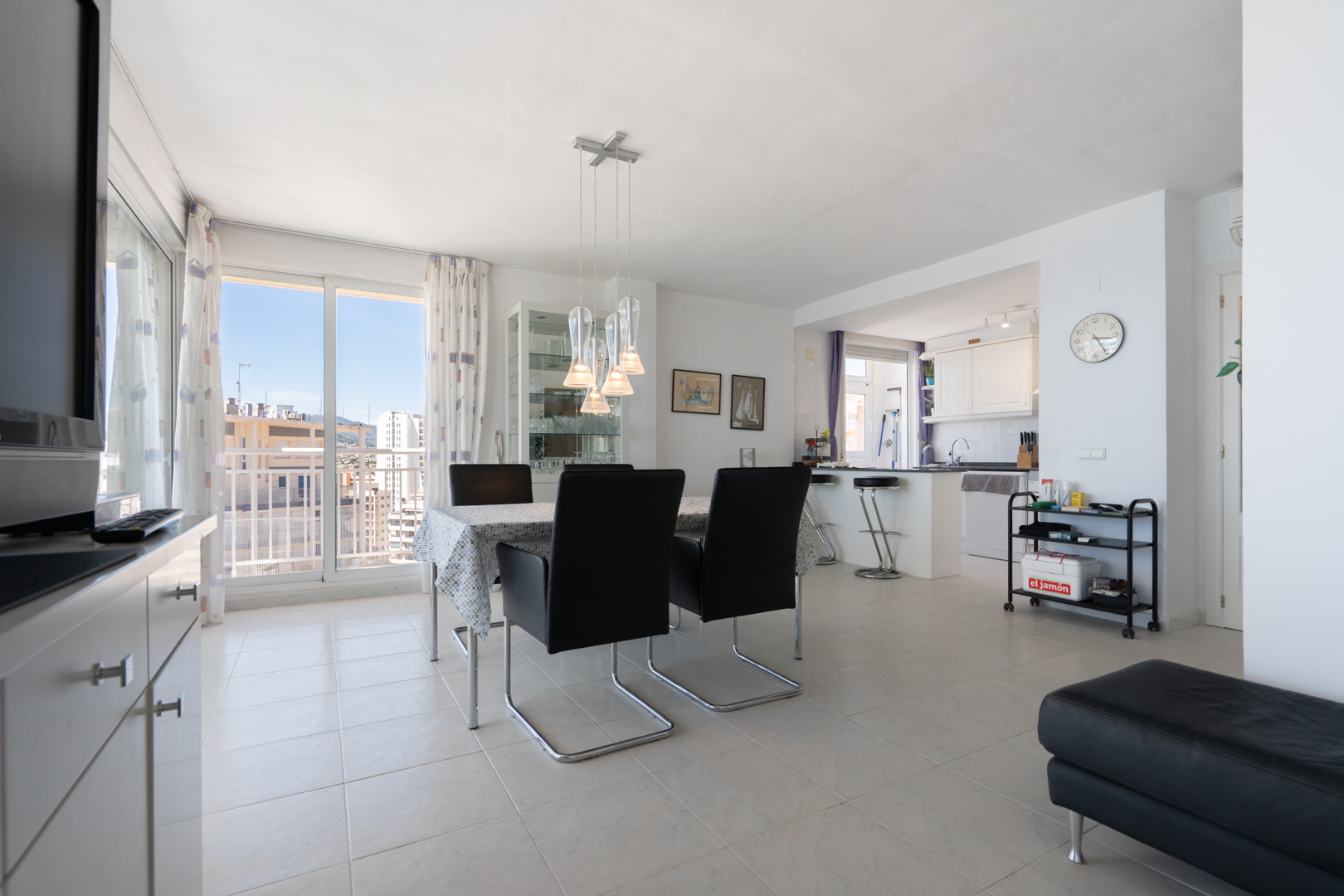 Lumineux salon-salle à manger ouvert avec suspension moderne, sol carrelé et accès balcon offrant des vues urbaines, attenant à une cuisine semi-ouverte.