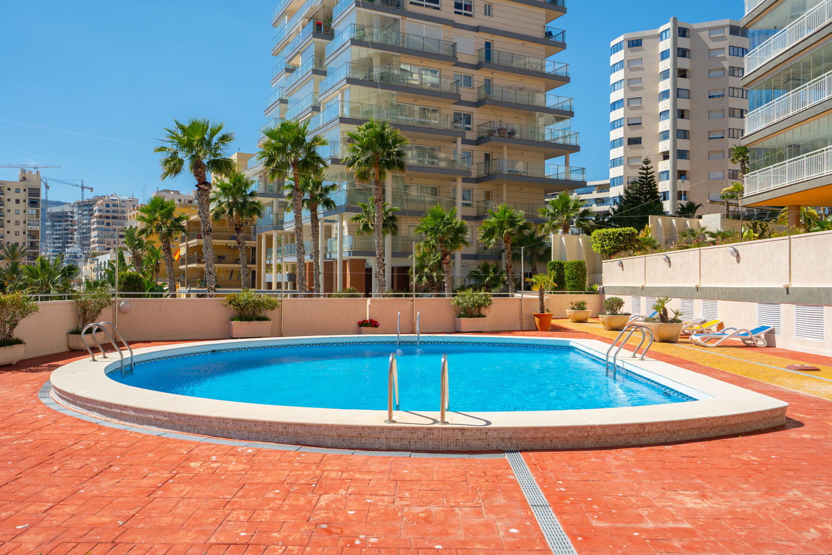 Piscine communautaire incurvée aux eaux bleues, entourée d'une terrasse carrelée rouge et jaune. Des palmiers et des immeubles résidentiels modernes aux balcons vitrés se profilent à l'arrière-plan.