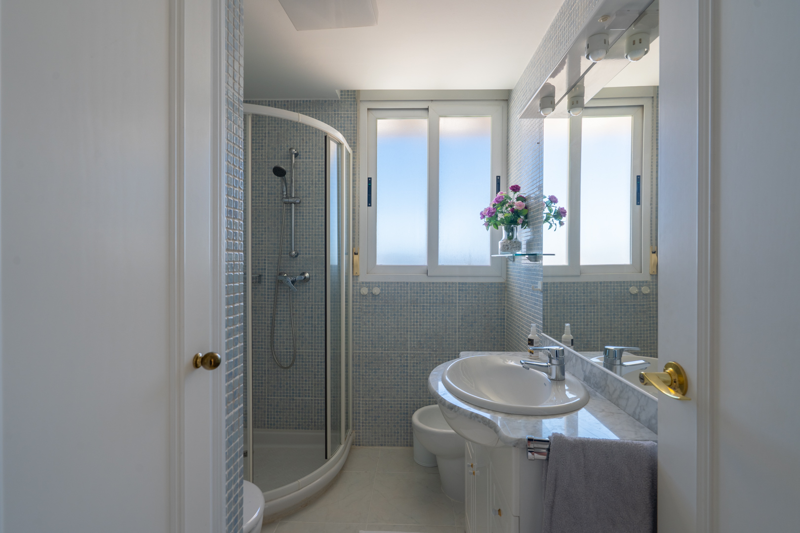 Salle de bain avec cabine de douche d'angle, meuble-lavabo blanc en marbre et carreaux de mosaïque bleu clair. Fenêtre et miroir éclairé par des spots.