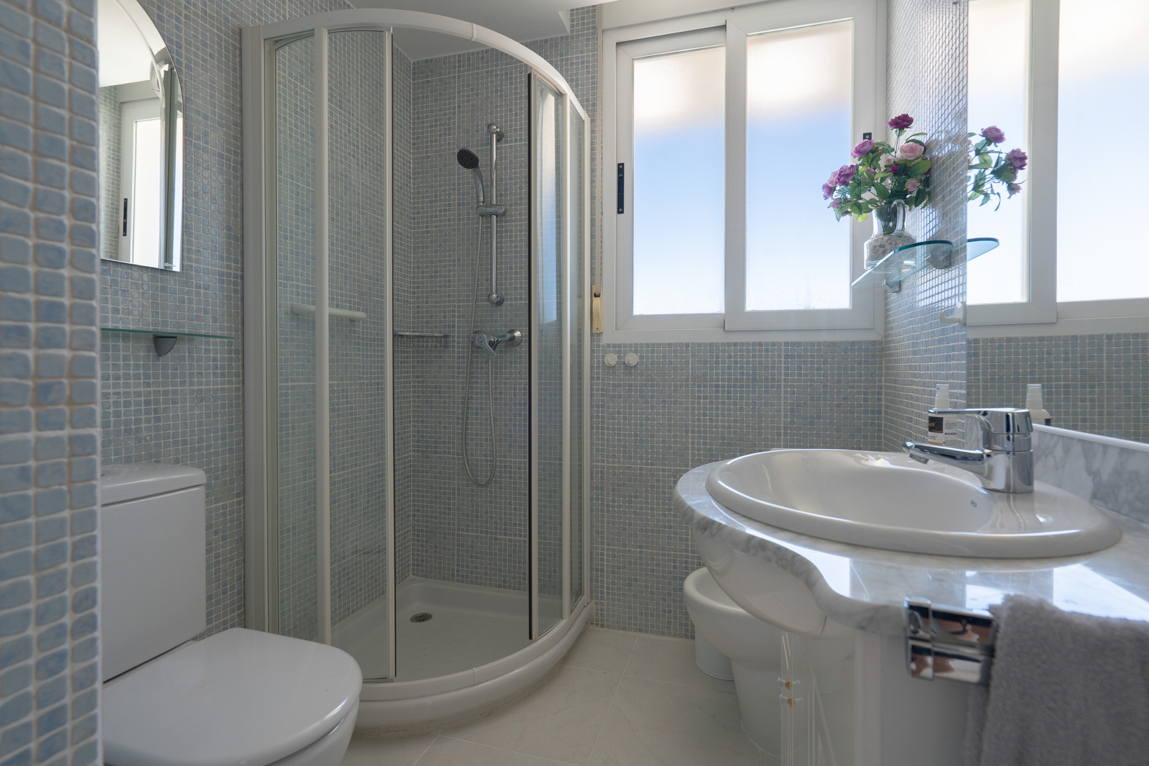 Salle de bain avec douche d'angle, toilettes, bidet et lavabo sur meuble effet marbre. Les murs sont recouverts de mosaïque bleu clair, éclairée par des fenêtres.