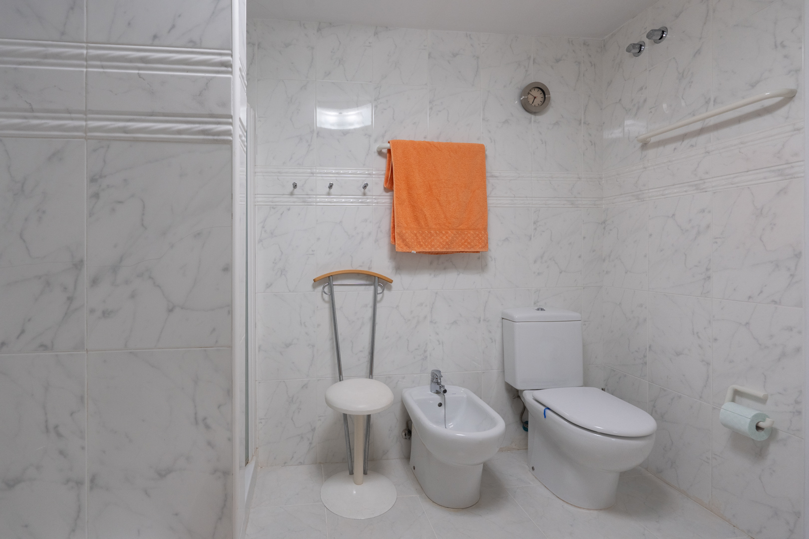 Salle de bain carrelée effet marbre blanc, comprenant des toilettes, un bidet et un espace douche fonctionnel avec barre d'appui. Un horloge murale et des patères sont également présents.