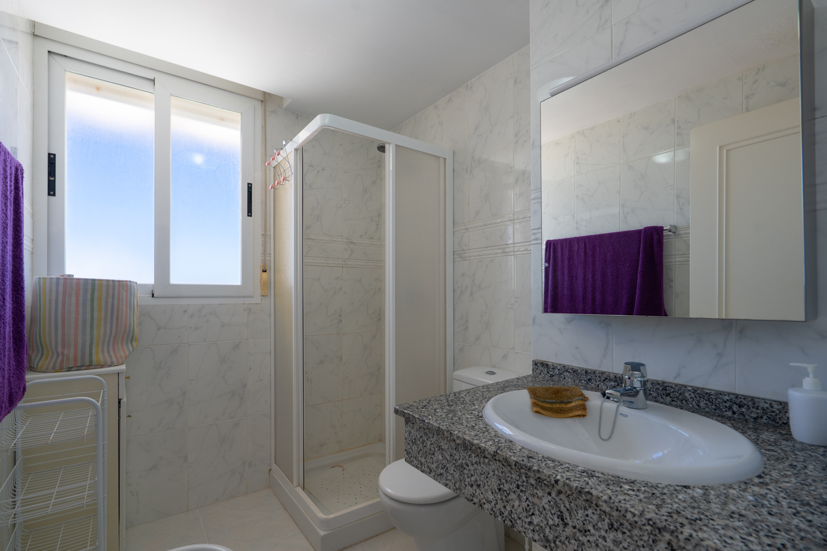 Salle de bain fonctionnelle avec carrelage mural effet marbre blanc, meuble-lavabo en granit moucheté et lavabo ovale. Comprend une cabine de douche d'angle et fenêtre.