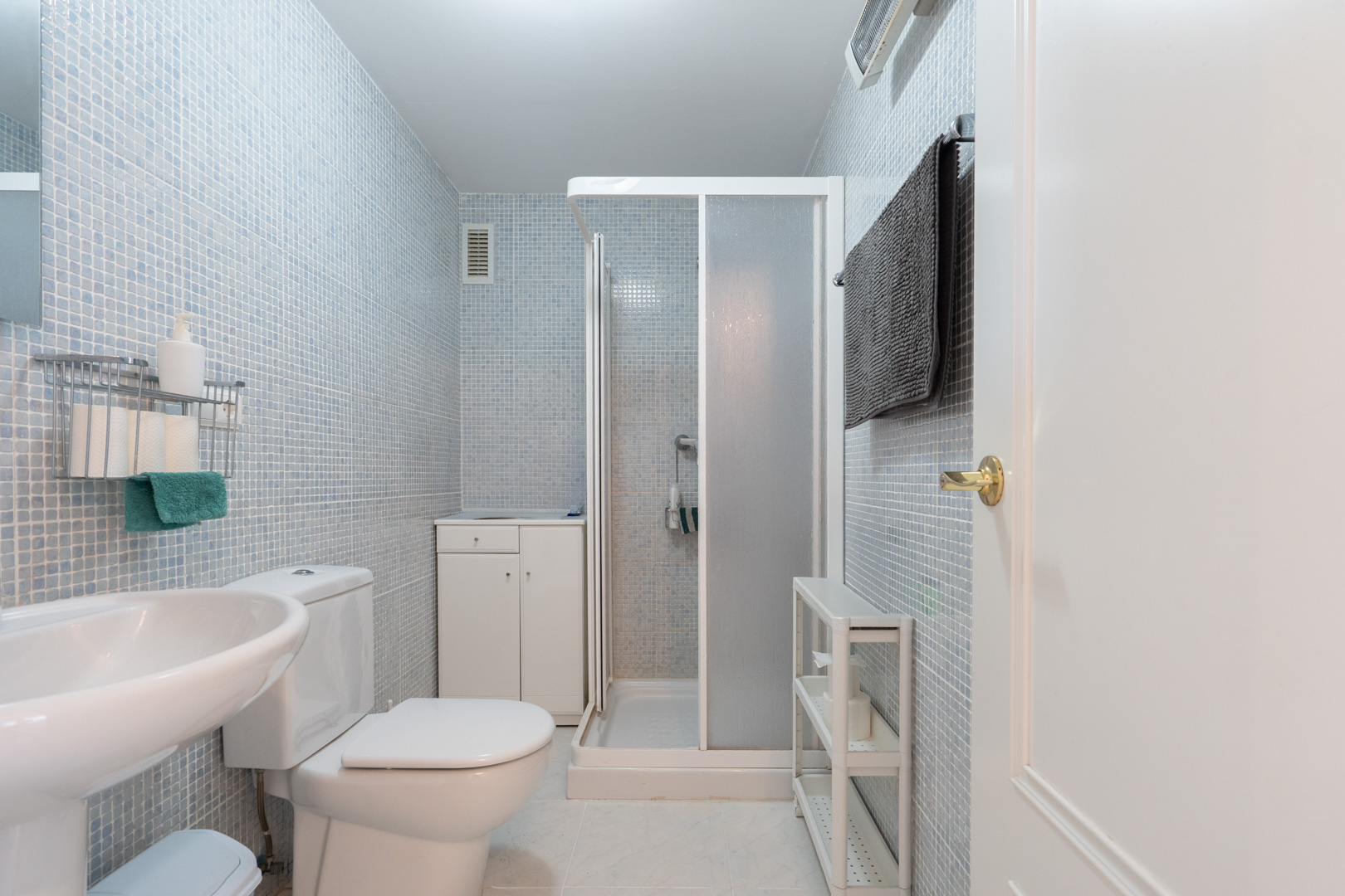 Salle de bain fonctionnelle avec murs en mosaïque bleu clair, équipée de toilettes, d'un lavabo sur pied et d'une cabine de douche. Comprend un petit meuble de rangement et des étagères.