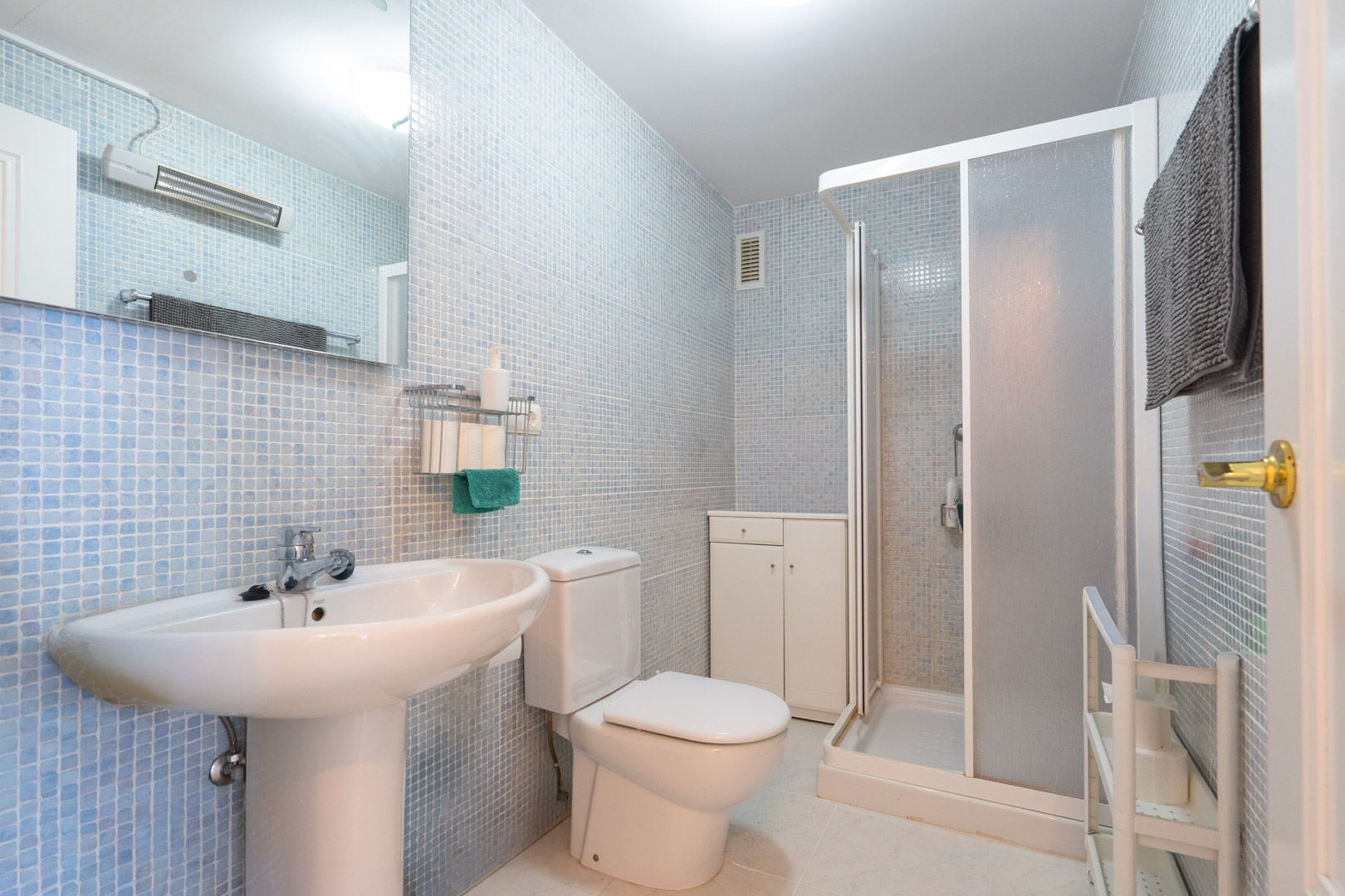 Salle de bain fonctionnelle avec murs en mosaïque bleu clair, lavabo sur pied, toilettes, cabine de douche vitrée dépolie et meuble de rangement blanc.