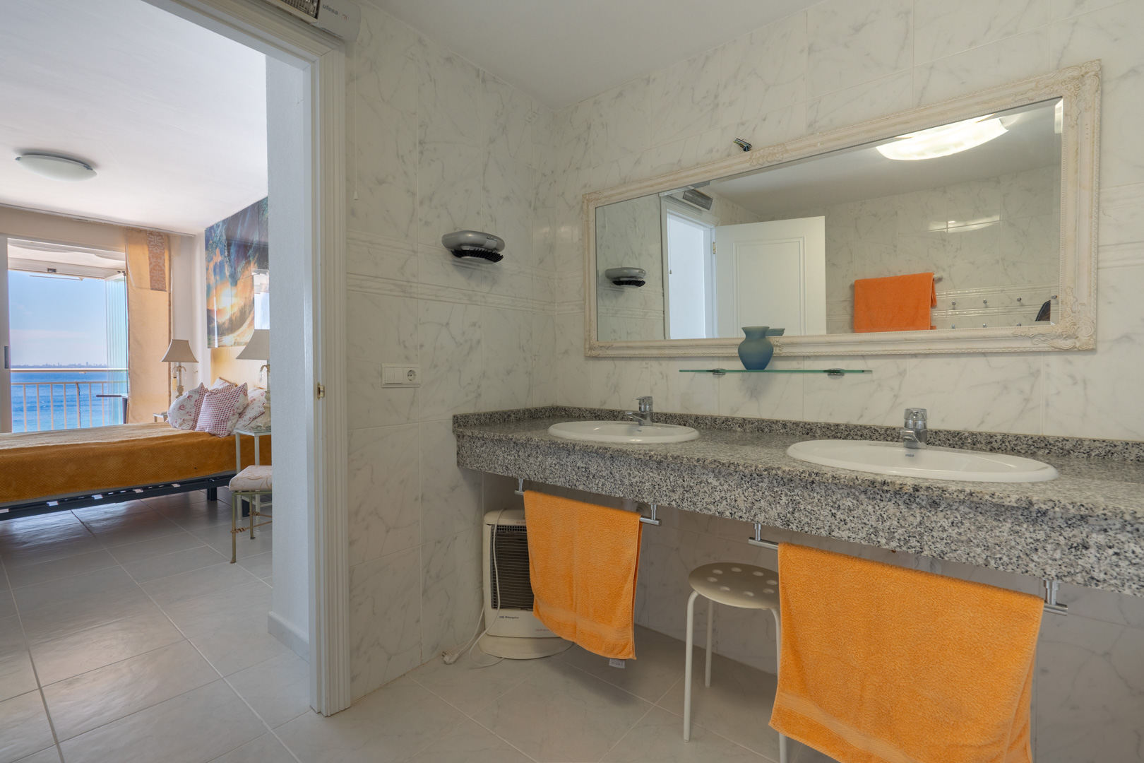 Salle de bain principale avec double vasque en granit, murs effet marbre et miroir orné, offrant une vue directe sur la chambre et la mer.