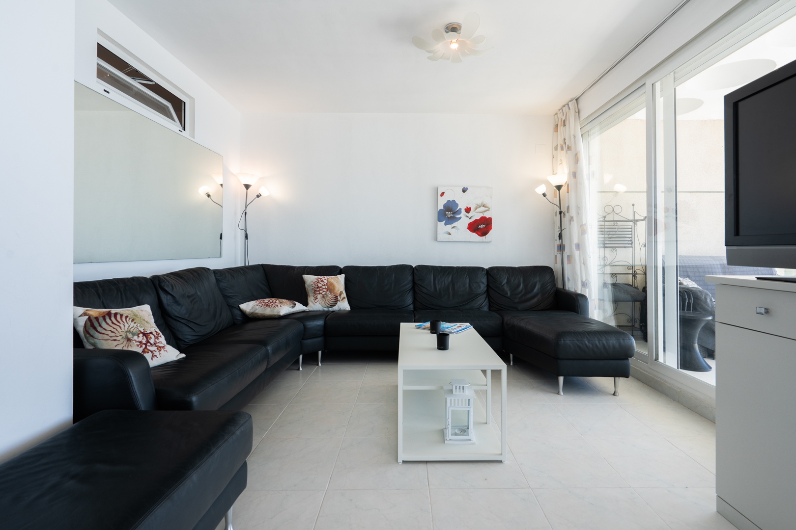 Salon spacieux avec un grand canapé d'angle noir et coussins décoratifs, sol carrelé blanc et table basse moderne. Les portes coulissantes vitrées s'ouvrent sur une terrasse.