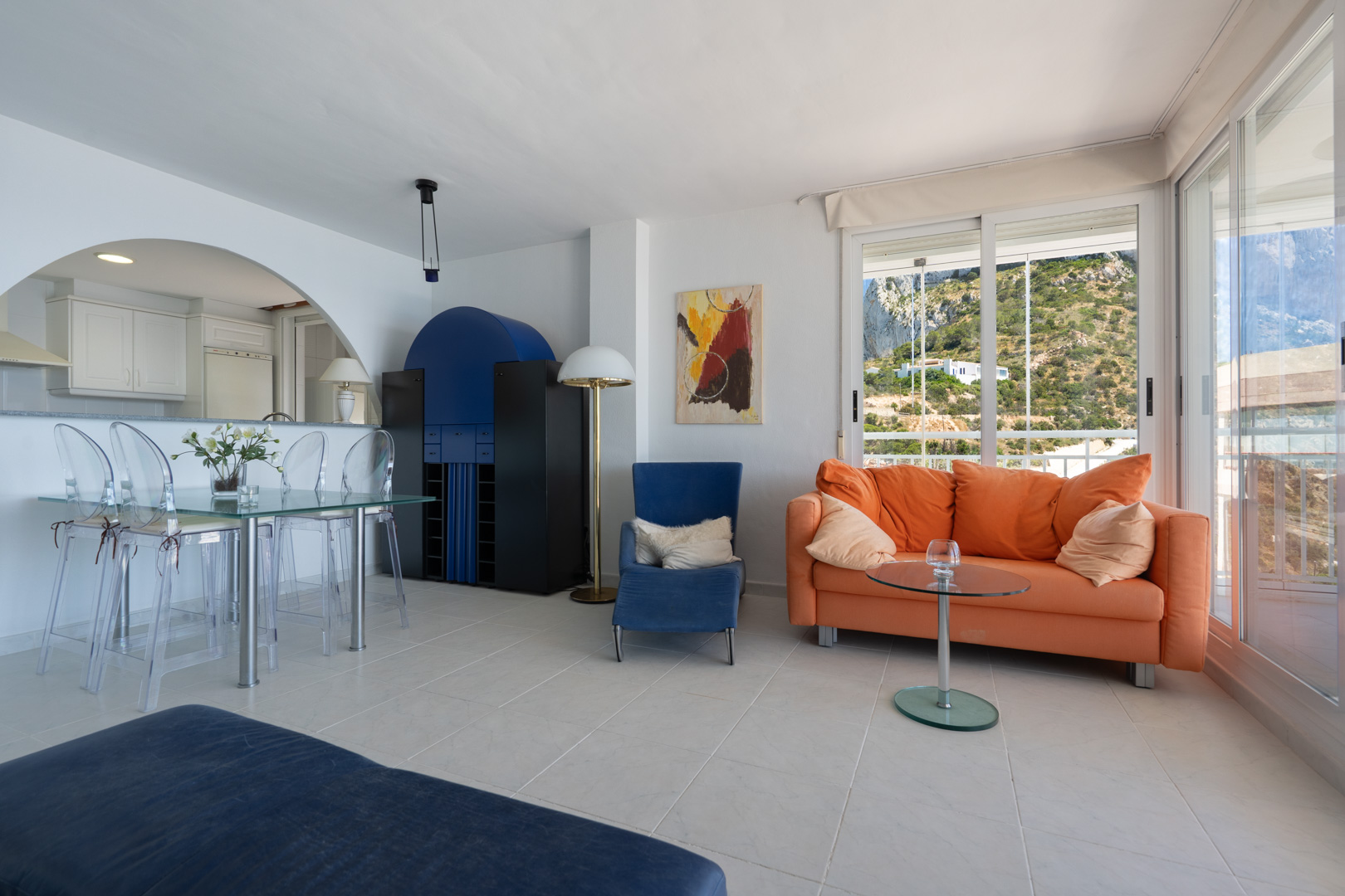 Séjour-salle à manger lumineux avec carrelage clair, table en verre et canapé orange. Grandes baies vitrées offrant une vue sur la colline verdoyante.
