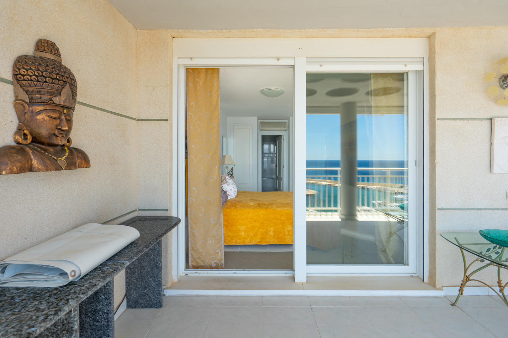 Terrasse couverte avec carrelage clair et murs texturés, donnant sur une chambre avec vue sur la mer via une porte coulissante en verre. Présente une console en granit et des éléments décoratifs.